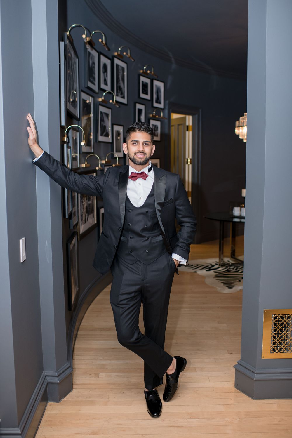 Elegant man in black tuxedo with burgundy bow tie posing in a sophisticated hallway with framed photos.
