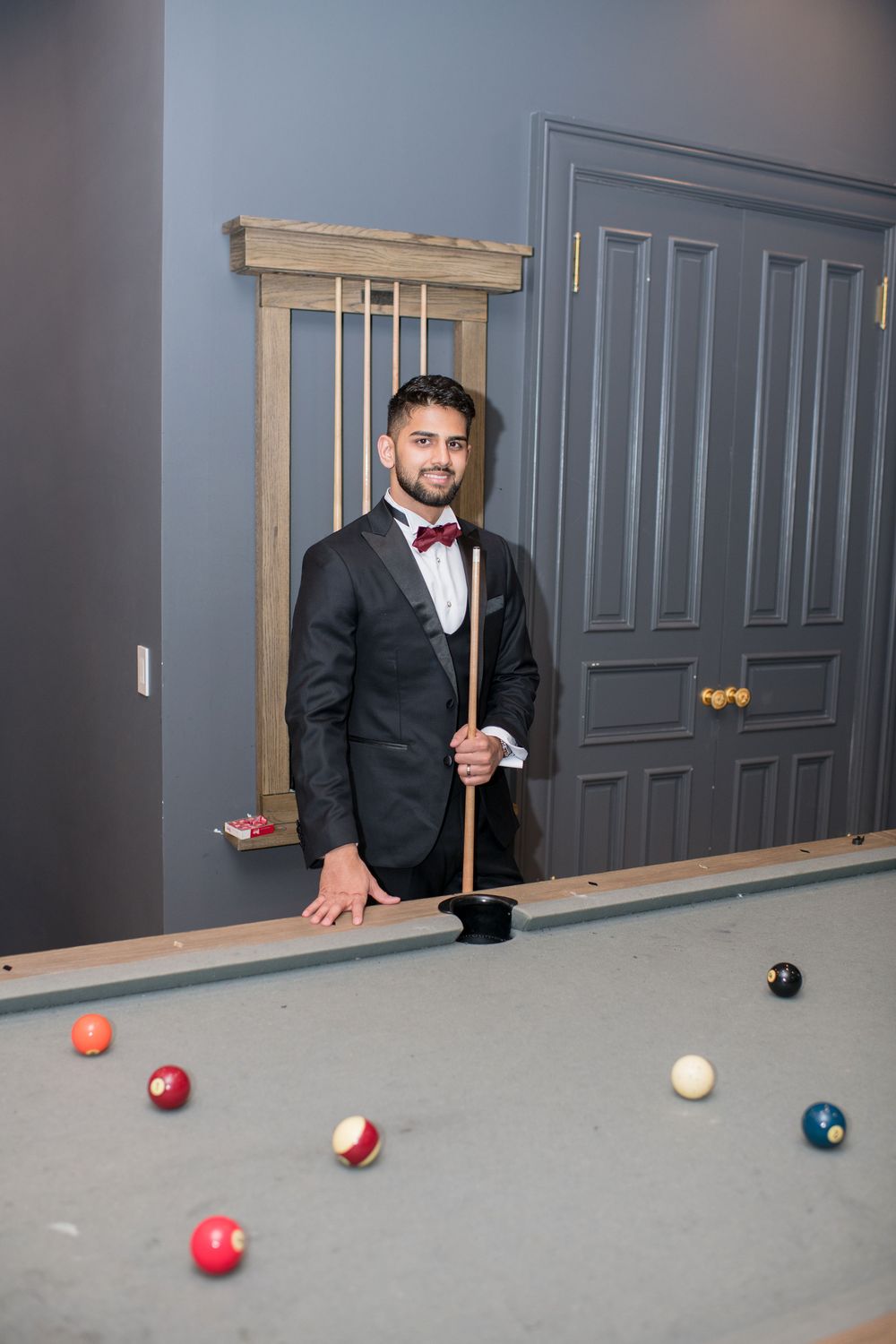 Smiling man in black tuxedo with red bow tie holding a pool cue beside a billiards table.