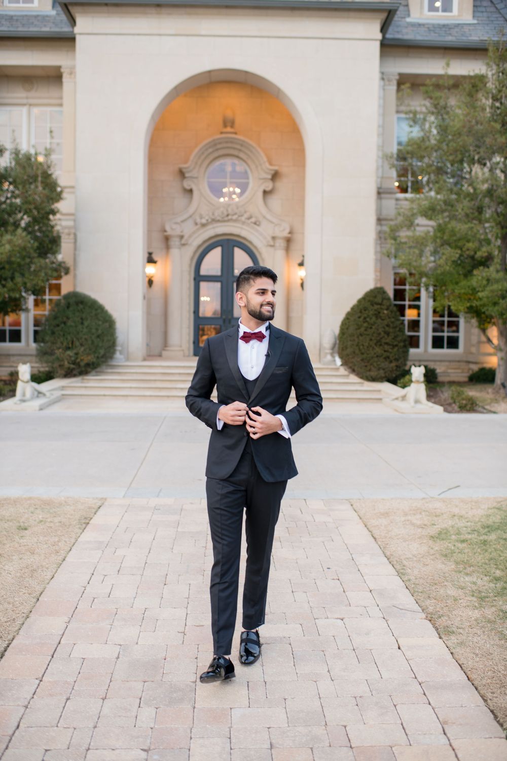 Dapper groom in black tuxedo with burgundy bow tie walks along brick path before elegant mansion venue.