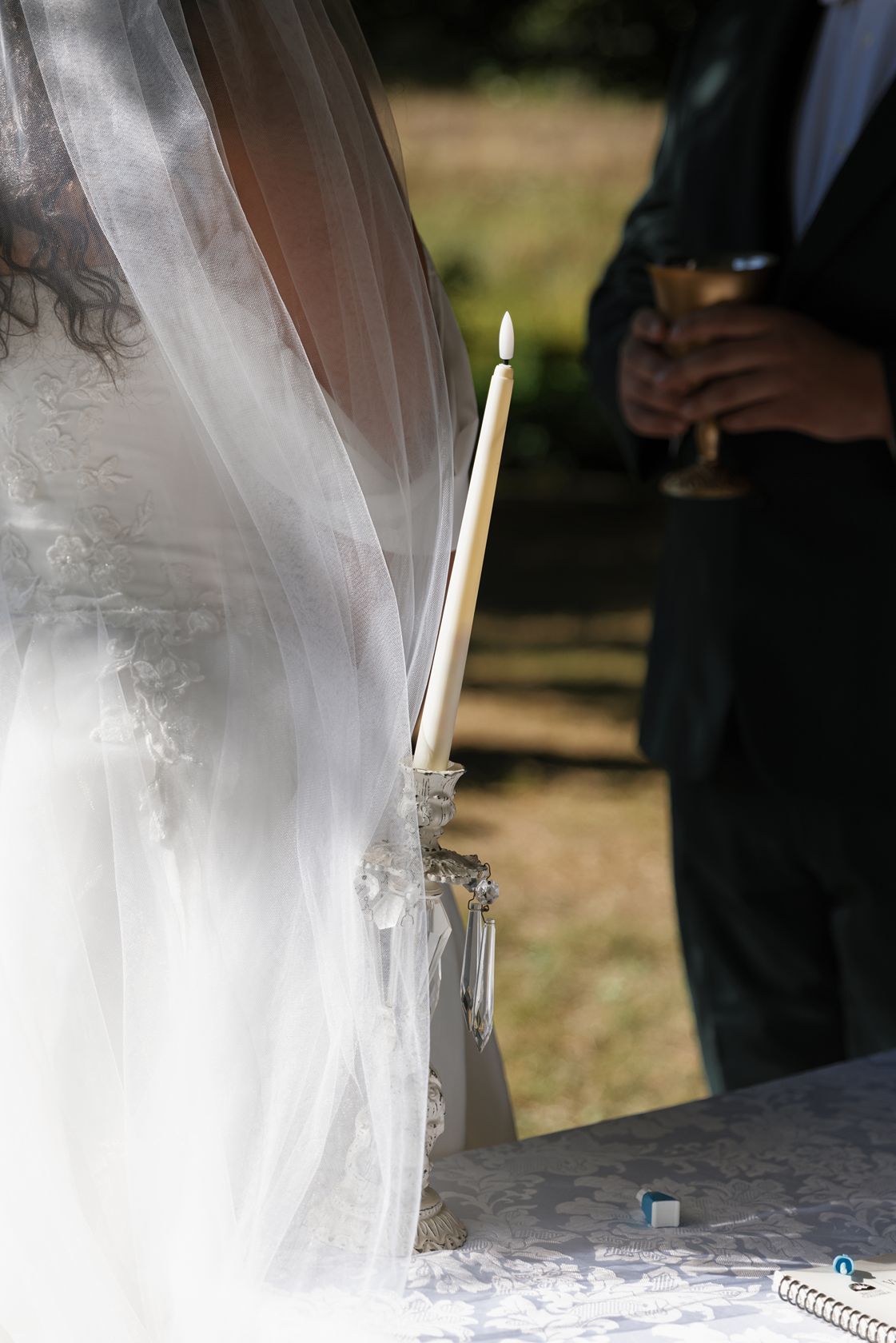 Elegant wedding candle in crystal holder beside bride's veil during outdoor ceremony.