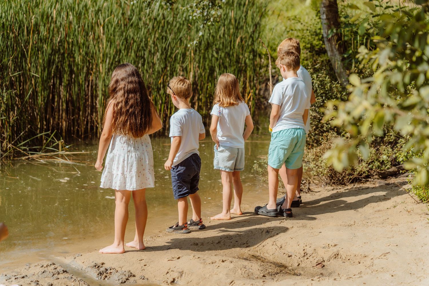 Four children standing barefoot on a sandy riverbank surrounded by lush green reeds and trees.