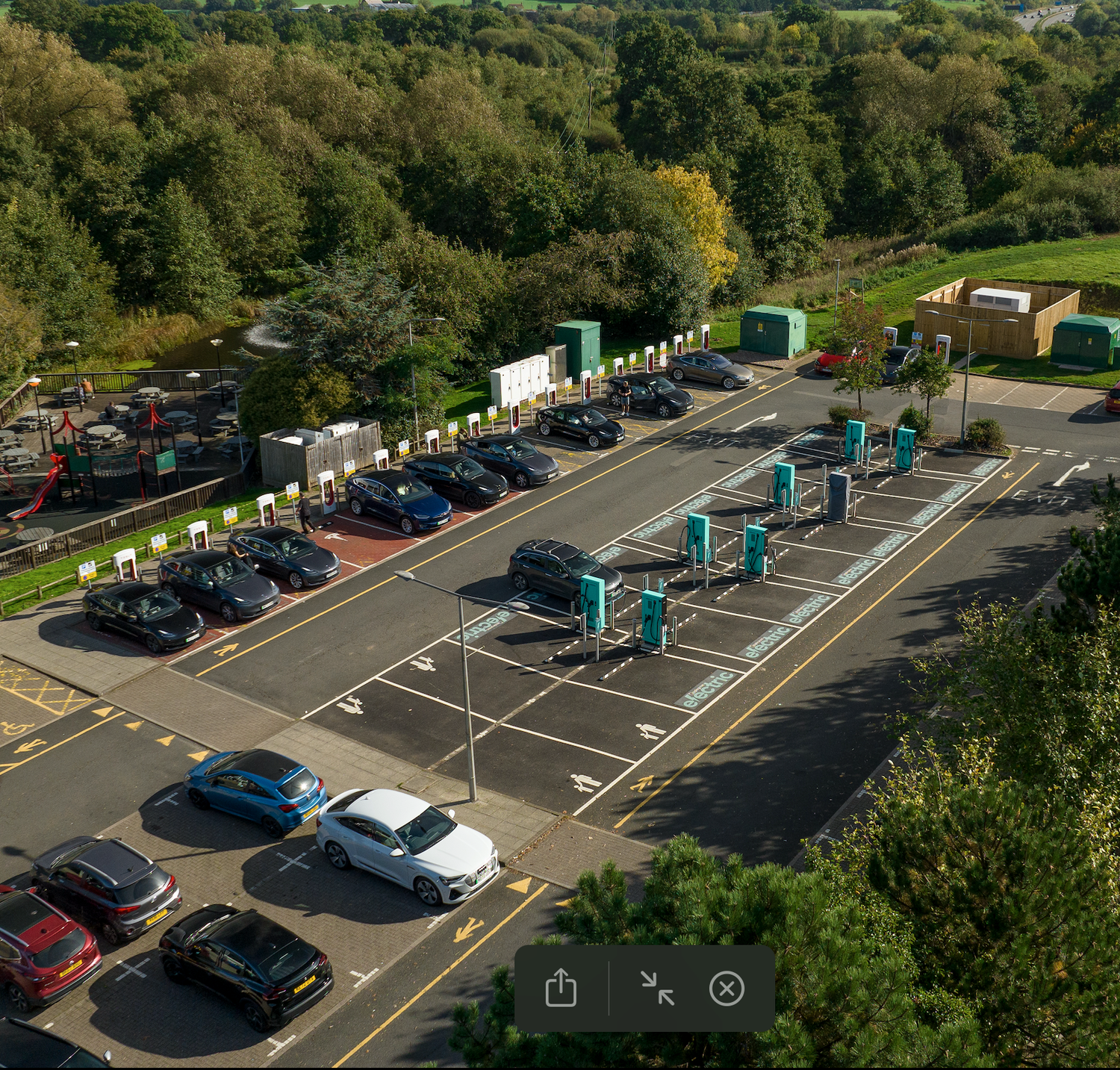 Aerial view of an electric vehicle charging station with multiple EV chargers in a parking lot surrounded by trees.
