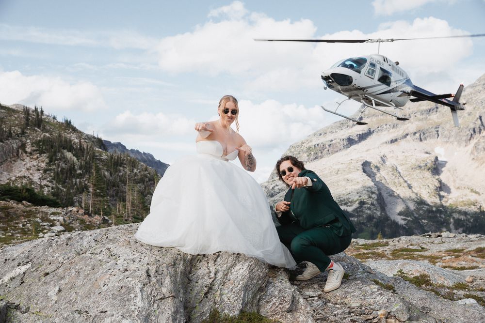 Bride in white gown and friend pose on mountain rocks with helicopter in background during elopement.
