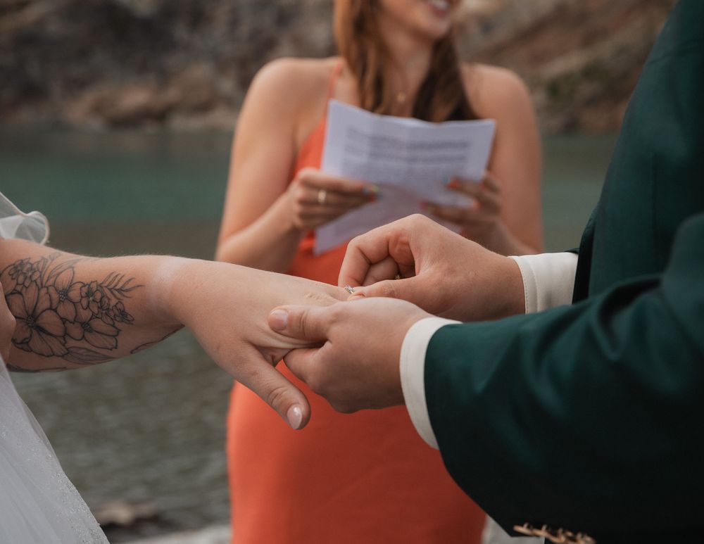 Close-up of ring exchange during wedding ceremony, bride with floral tattoo, officiant in orange dress.