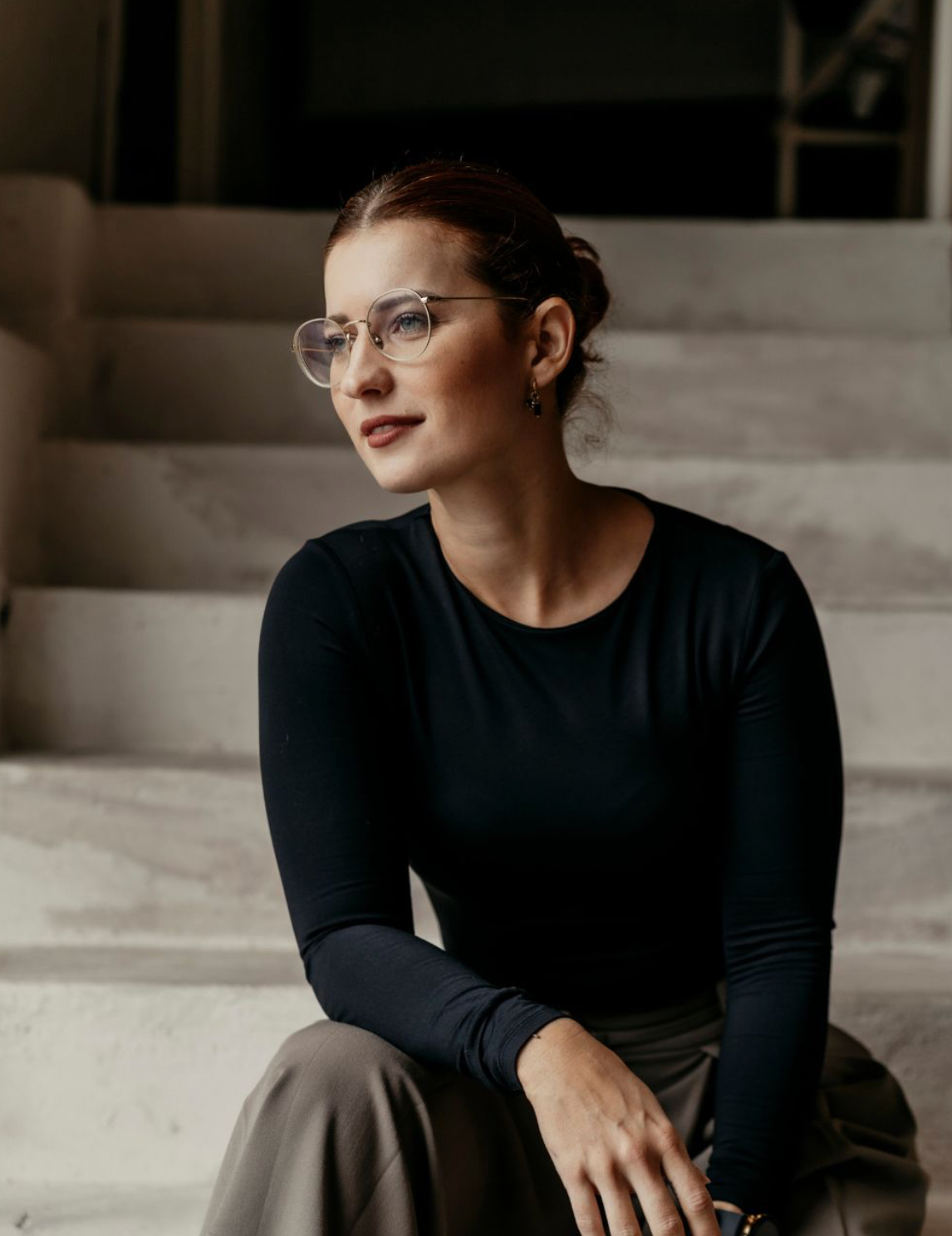 Woman with glasses and dark hair in a bun sits on steps wearing a black long-sleeve top and khaki pants.