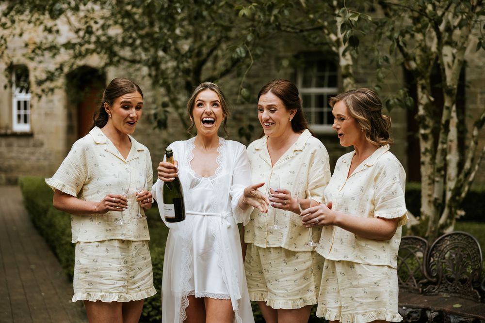 Bride in white robe laughing with three bridesmaids in matching cream sets, celebrating with champagne outdoors.