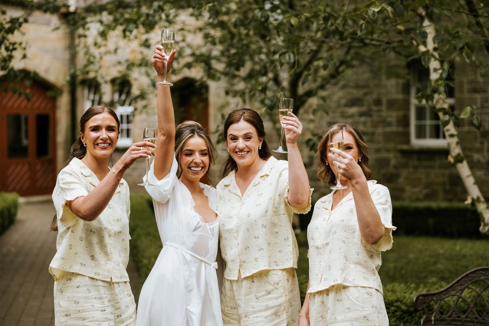 Four women in matching cream outfits raise champagne glasses outdoors at a wedding celebration.