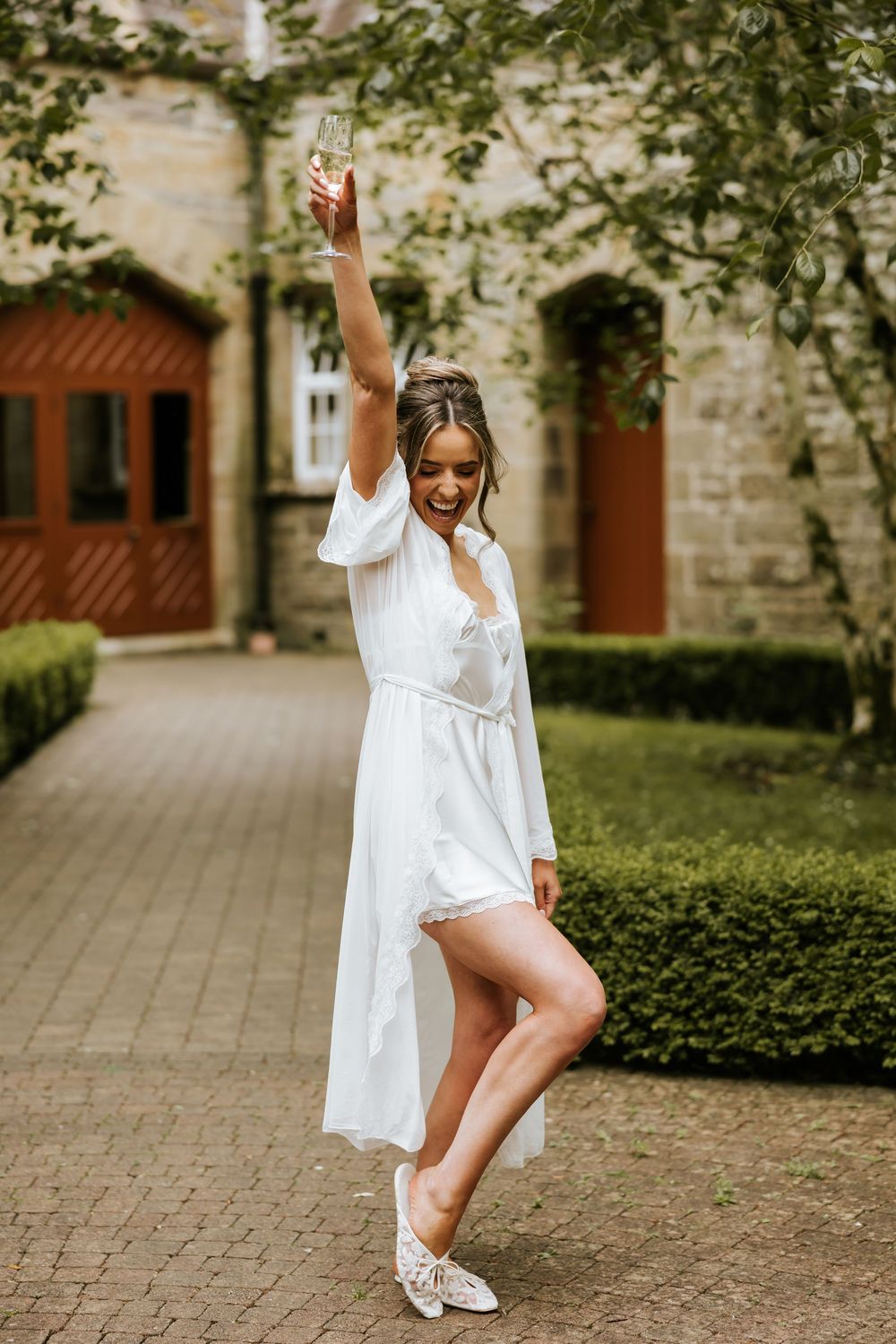 Bride in white robe raises champagne glass outdoors near stone building with lush greenery.