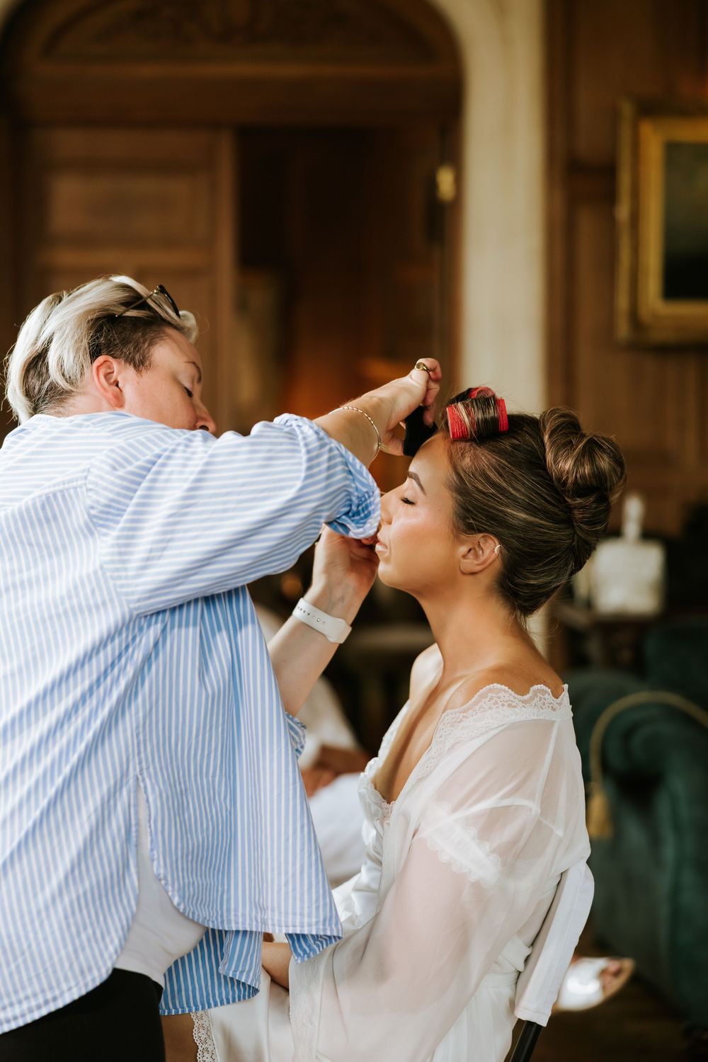 Makeup artist applying cosmetics to a bride with hair rollers in an elegant room.