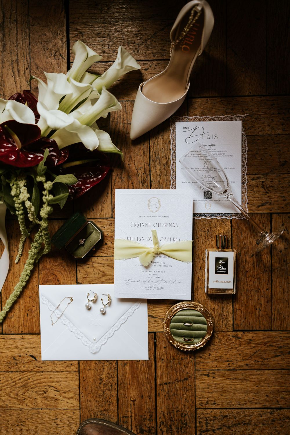 Elegant wedding flat lay with floral bouquet, invitation suite, pearl earrings, perfume, and nude heels on wood floor.