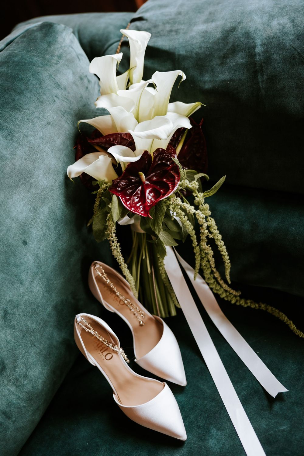 White bridal heels and calla lily bouquet with red anthuriums on a teal velvet sofa.