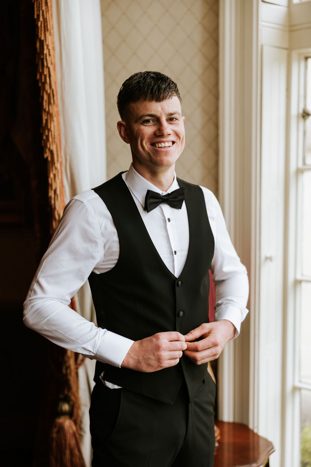 Smiling groom in black tuxedo vest and bow tie buttoning his waistcoat while standing near a window.