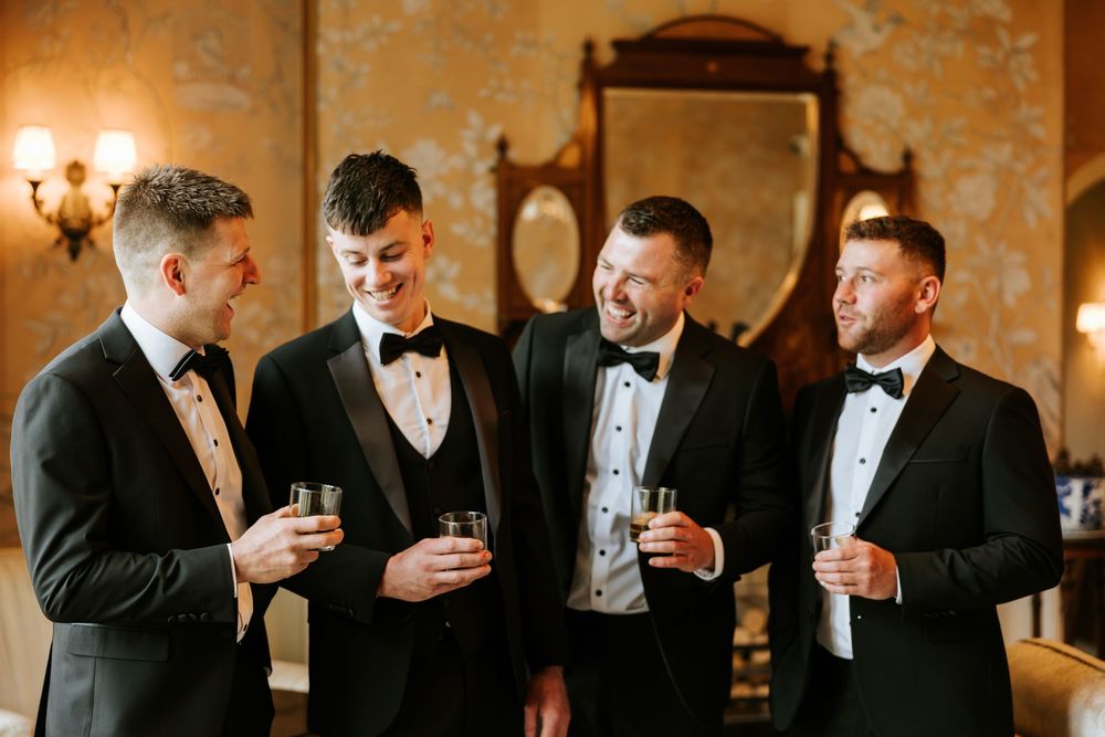 Four groomsmen in black tuxedos with bow ties toasting with drinks in an elegant vintage-styled room.