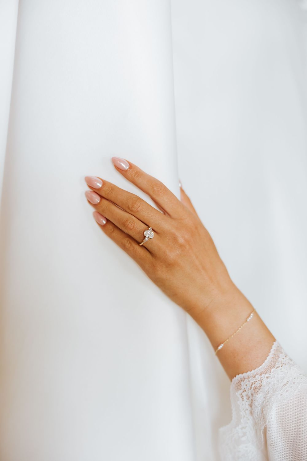 Close-up of bride's hand wearing a diamond solitaire engagement ring and delicate gold bracelet on white dress.