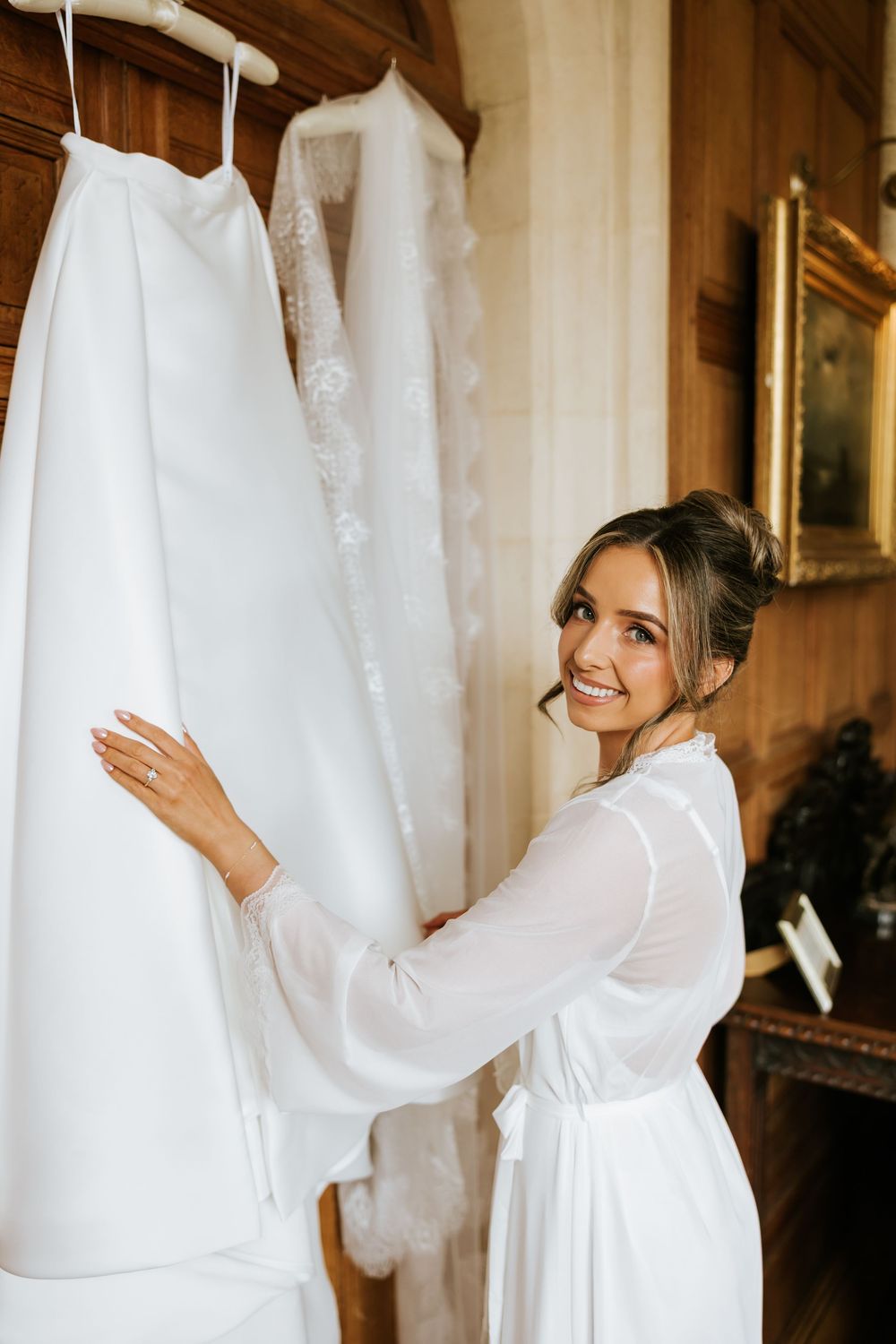Smiling bride in white robe admiring her wedding dress hanging in an elegant wood-paneled room.