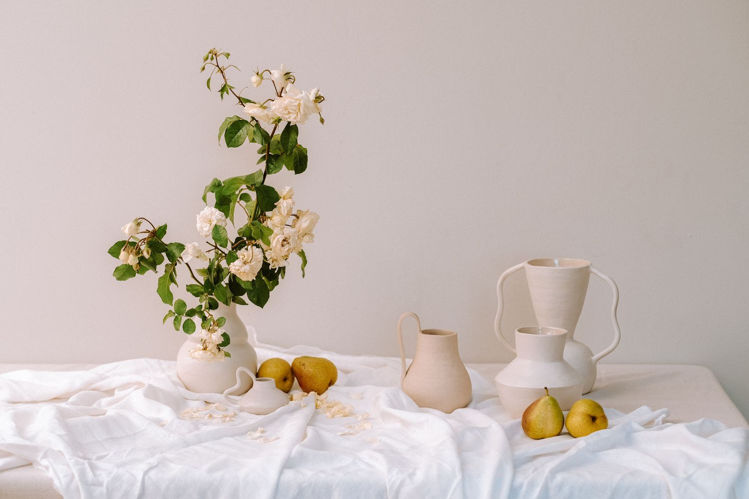 Elegant still life arrangement with white flowers, pears, and ceramic vases on a white linen-draped table.