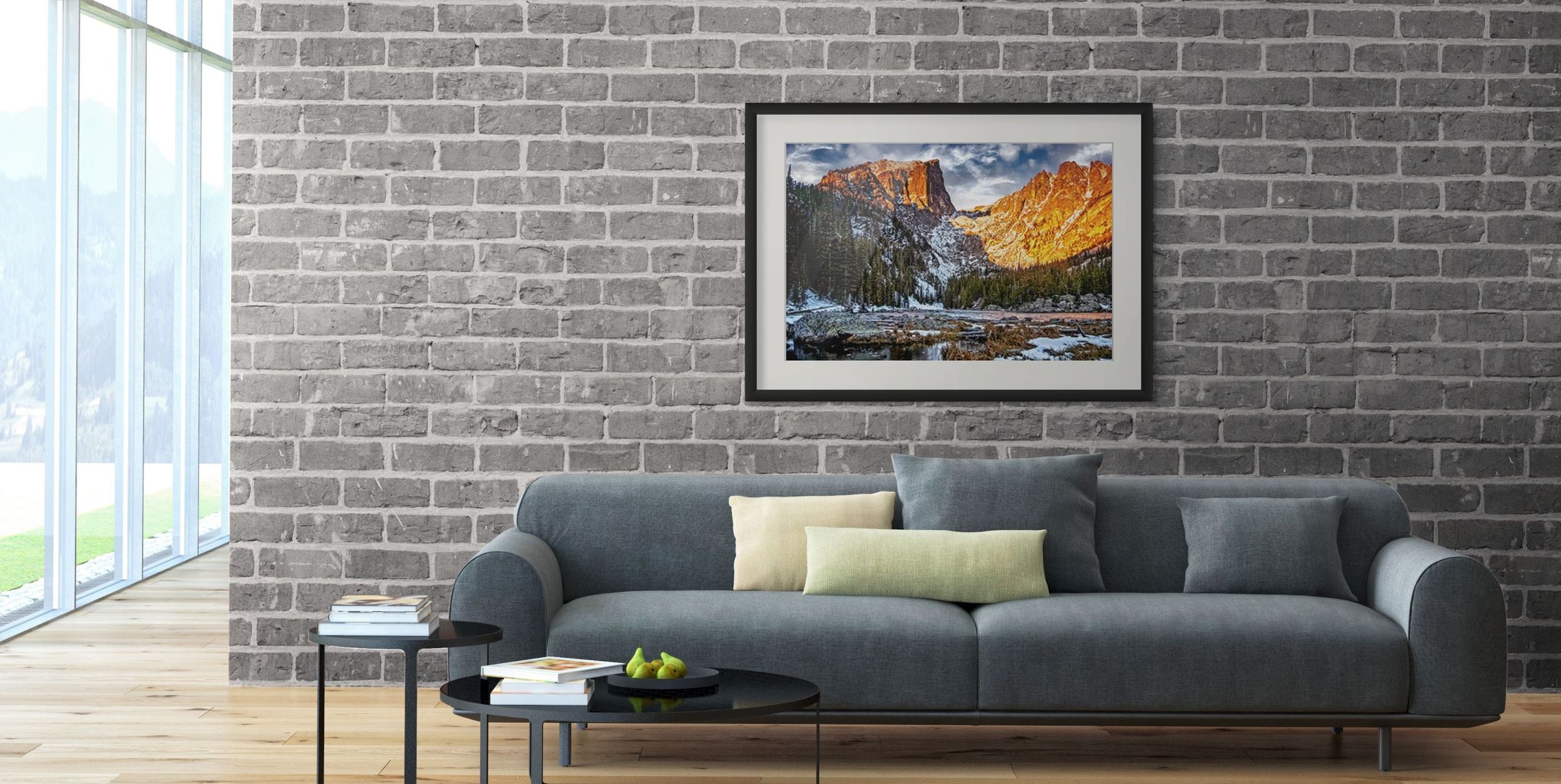 Framed landscape print of mountain and lake scene hanging on gray brick wall above modern gray sofa in contemporary living room.