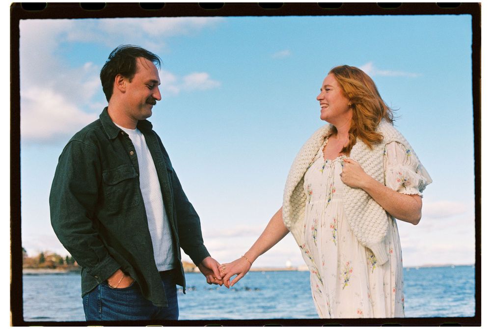 Couple holding hands and smiling at each other by the sea in front of blue skies on color film during a maternity photoshoot in Maine.