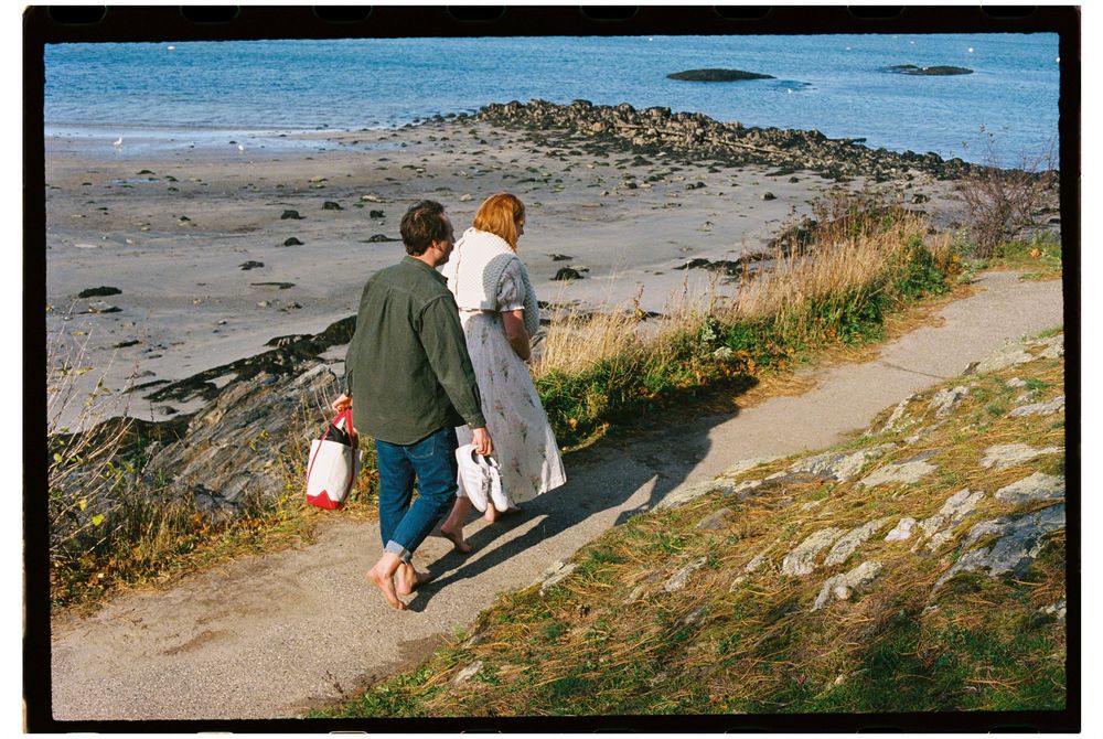 Couple walk together above the beach during a sunny day shoot celebrating their expected child during a maternity photoshoot on film.