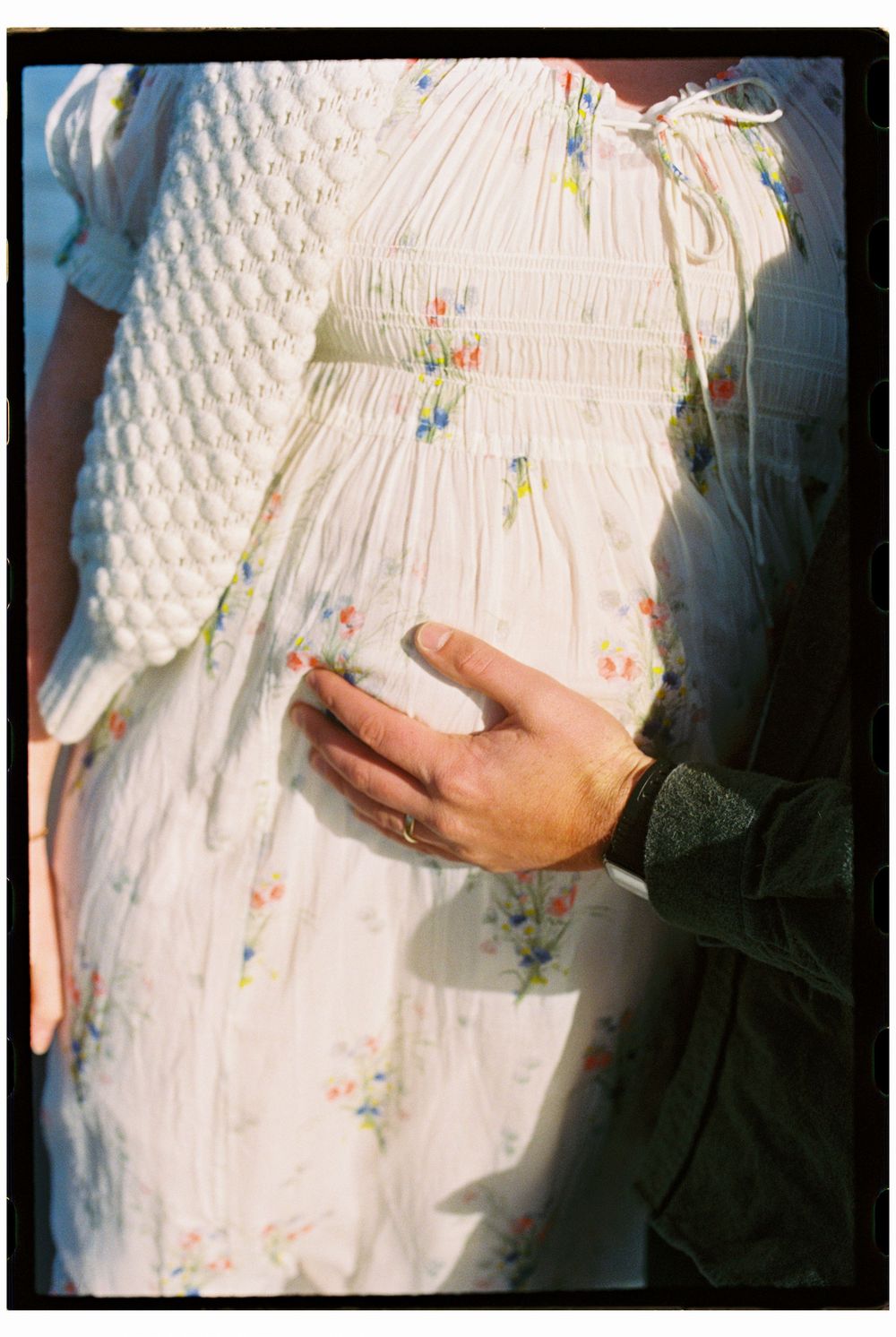 Detail photograph of a husband lovingly touching his wife's pregnant belly during a maternity photoshoot on film at Willard beach in Maine.