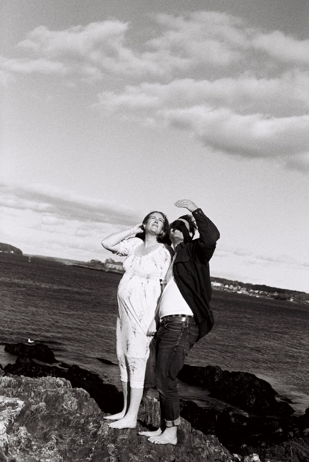 Couple stands together on the rocks by the ocean looking up together at the sky on black and white film during a maternity photoshoot.