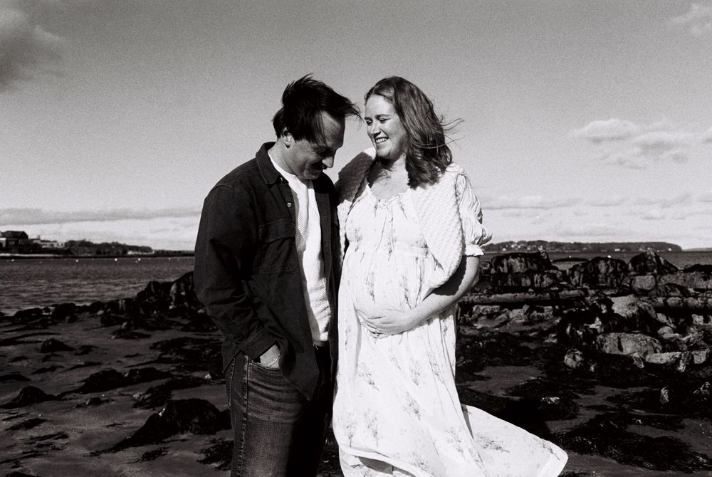 Maternity photoshoot on the beach in the sunshine on black and white film showing a couple close to each other while she holds her belly.