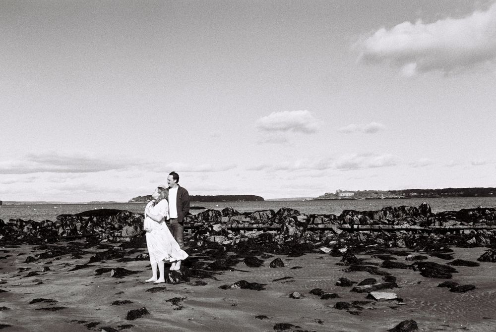 Couple expecting a baby pose together on the beach in a black and white film photo during an early winter maternity shoot in South Portland.