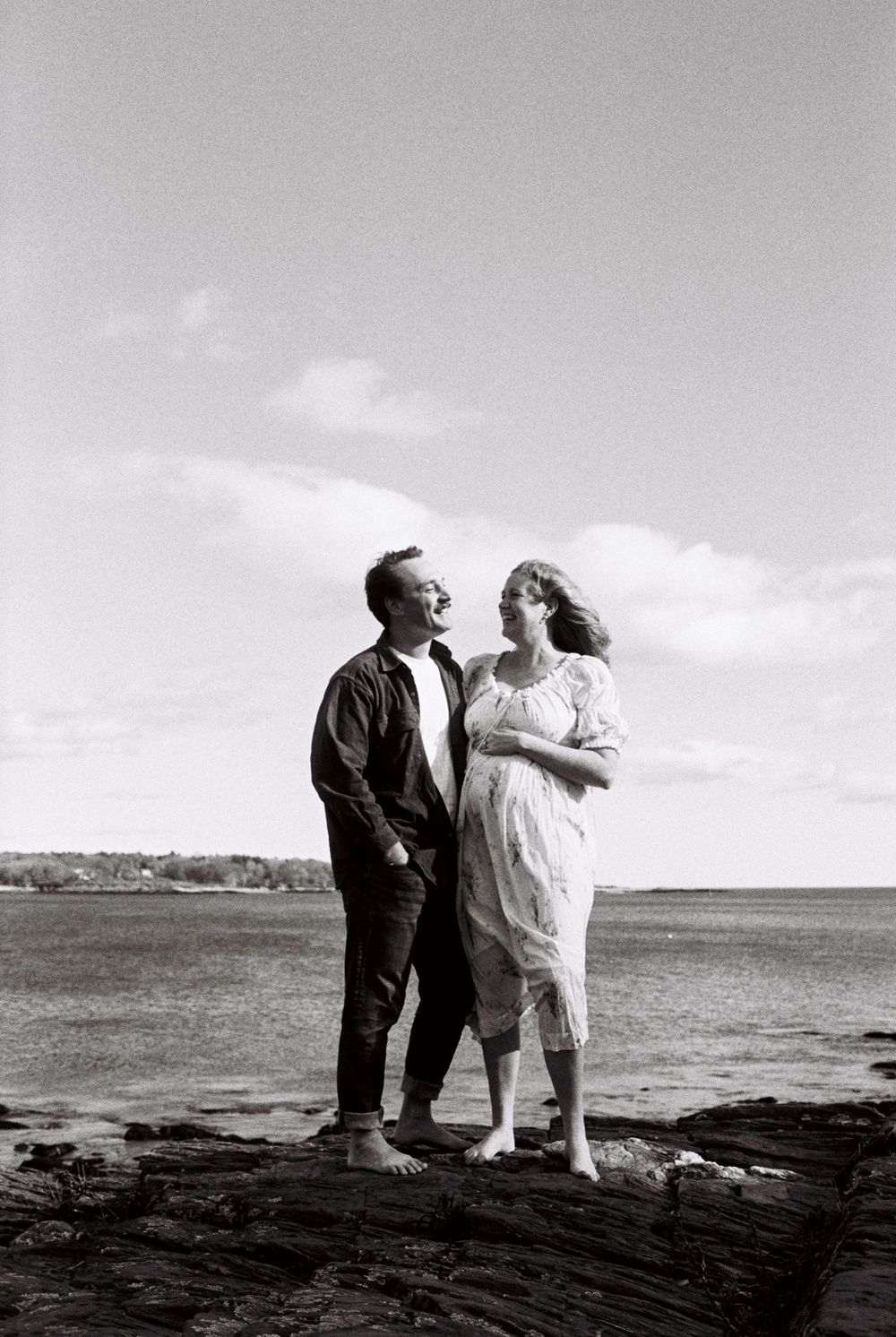 Black and white film photo of couple smiling and laughing by the ocean on the beach with her hand on her belly at Willard beach in Maine.