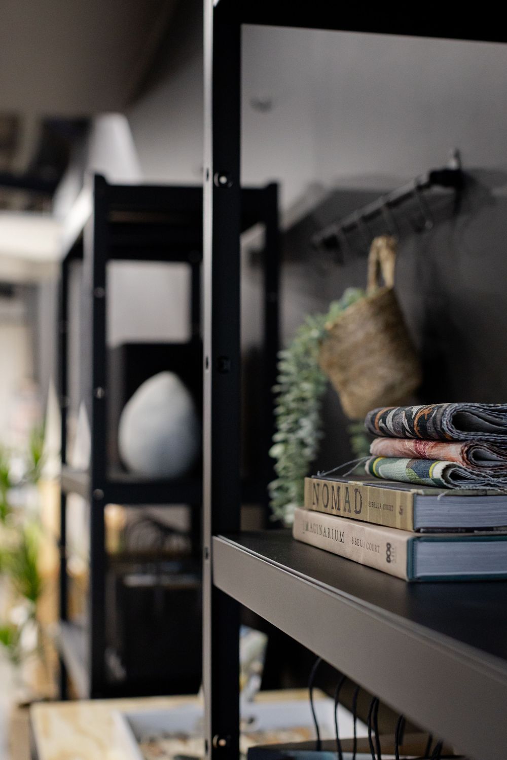 Black metal shelving unit displaying stacked books including Nomad title, decorative greenery, and woven basket in modern interior.