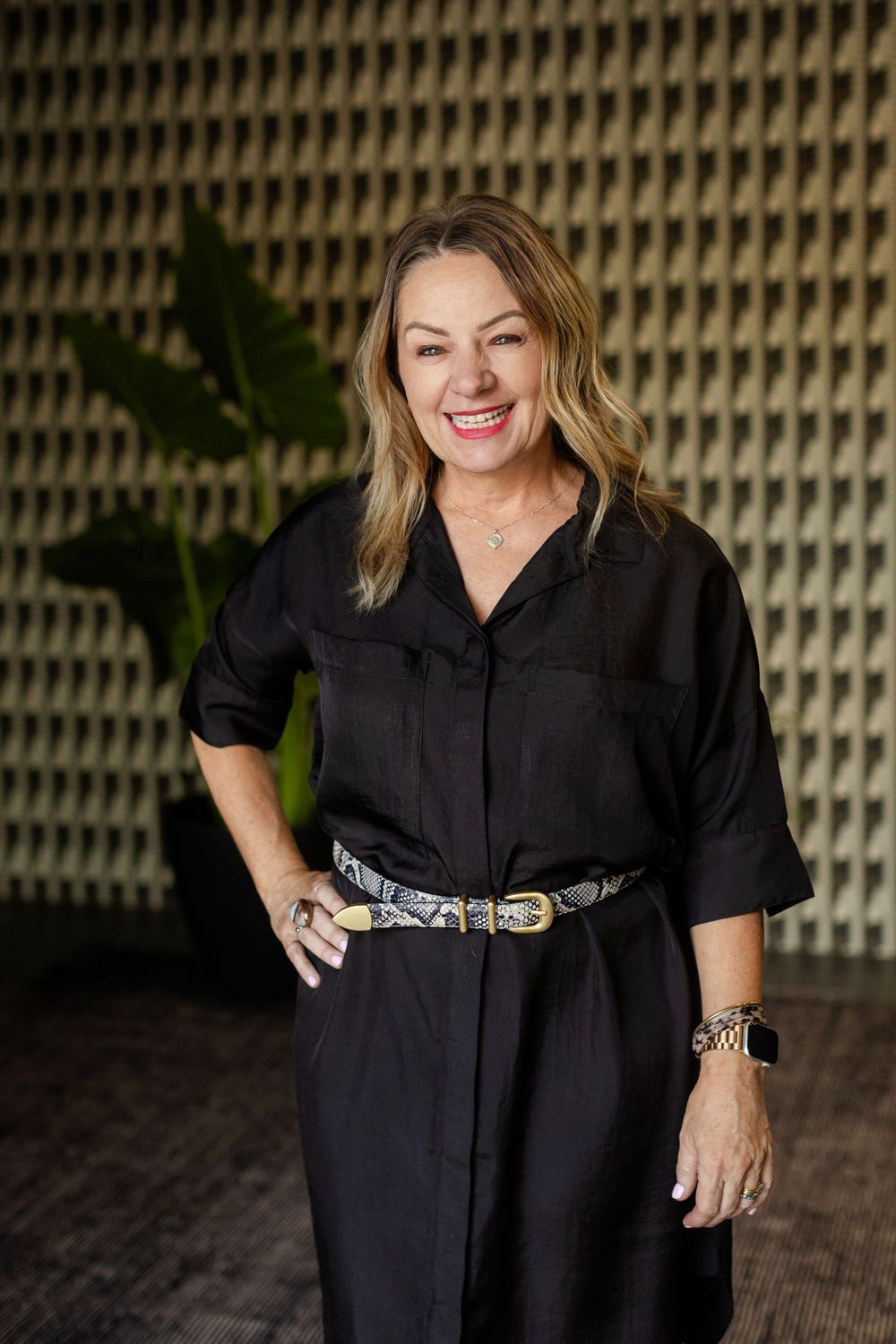 Smiling woman in black dress with decorative belt poses confidently in modern interior space.