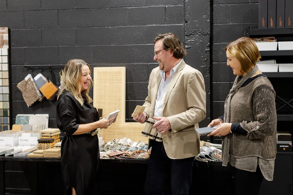 Three people conversing at a display table with fabric and material samples in a dark-walled showroom.