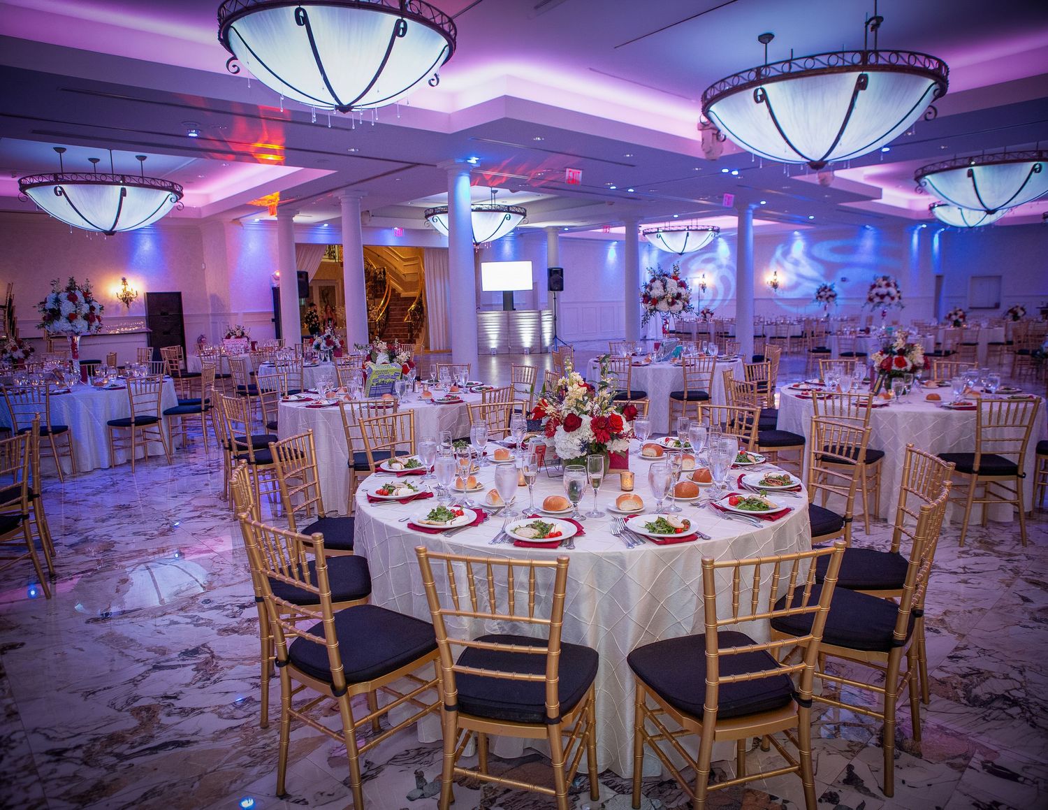 Elegant banquet hall with gold Chiavari chairs, round tables set for a wedding reception, purple uplighting, and floral centerpieces.
