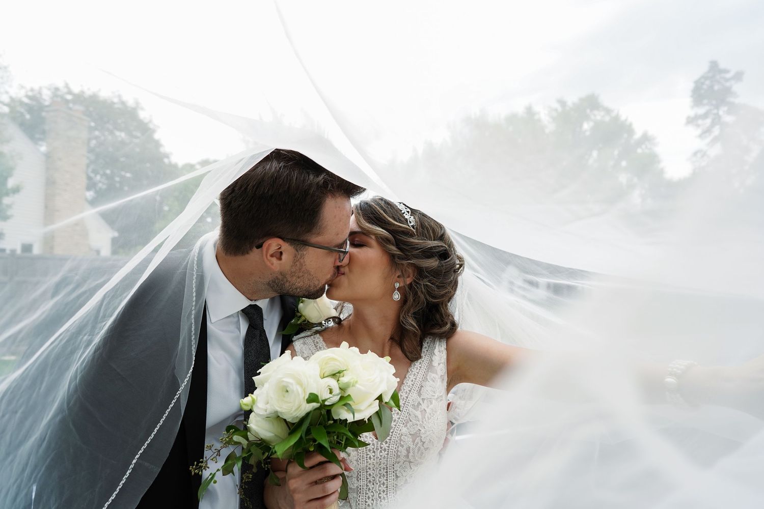Bride and groom kissing under flowing white veil while holding white rose bouquet.