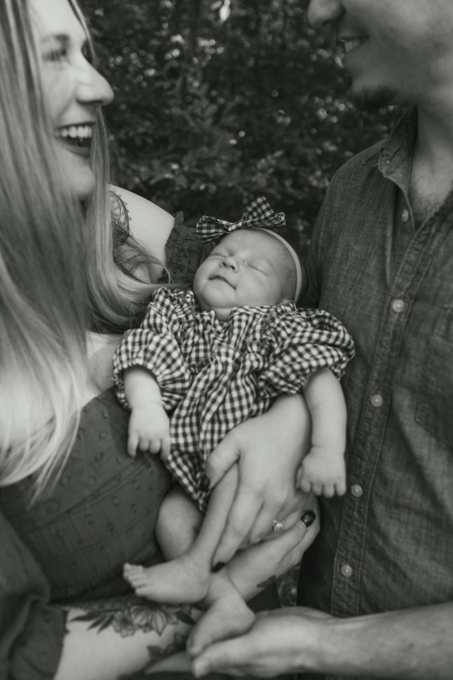 Black and white photo of smiling parents holding their newborn baby dressed in plaid outfit with bow headband outdoors.