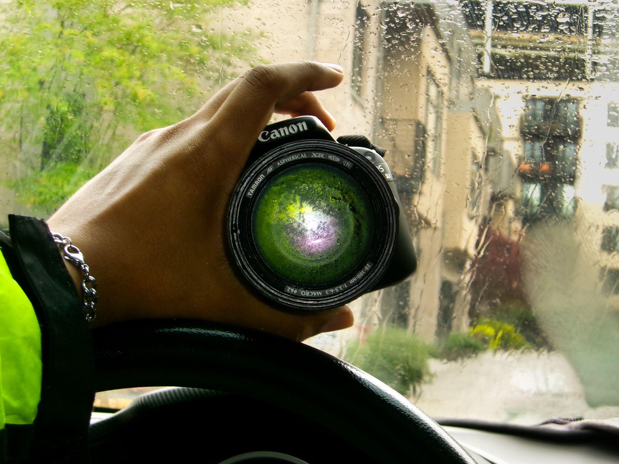 Hand holding Canon camera lens reflecting green foliage through rain-covered car window.
