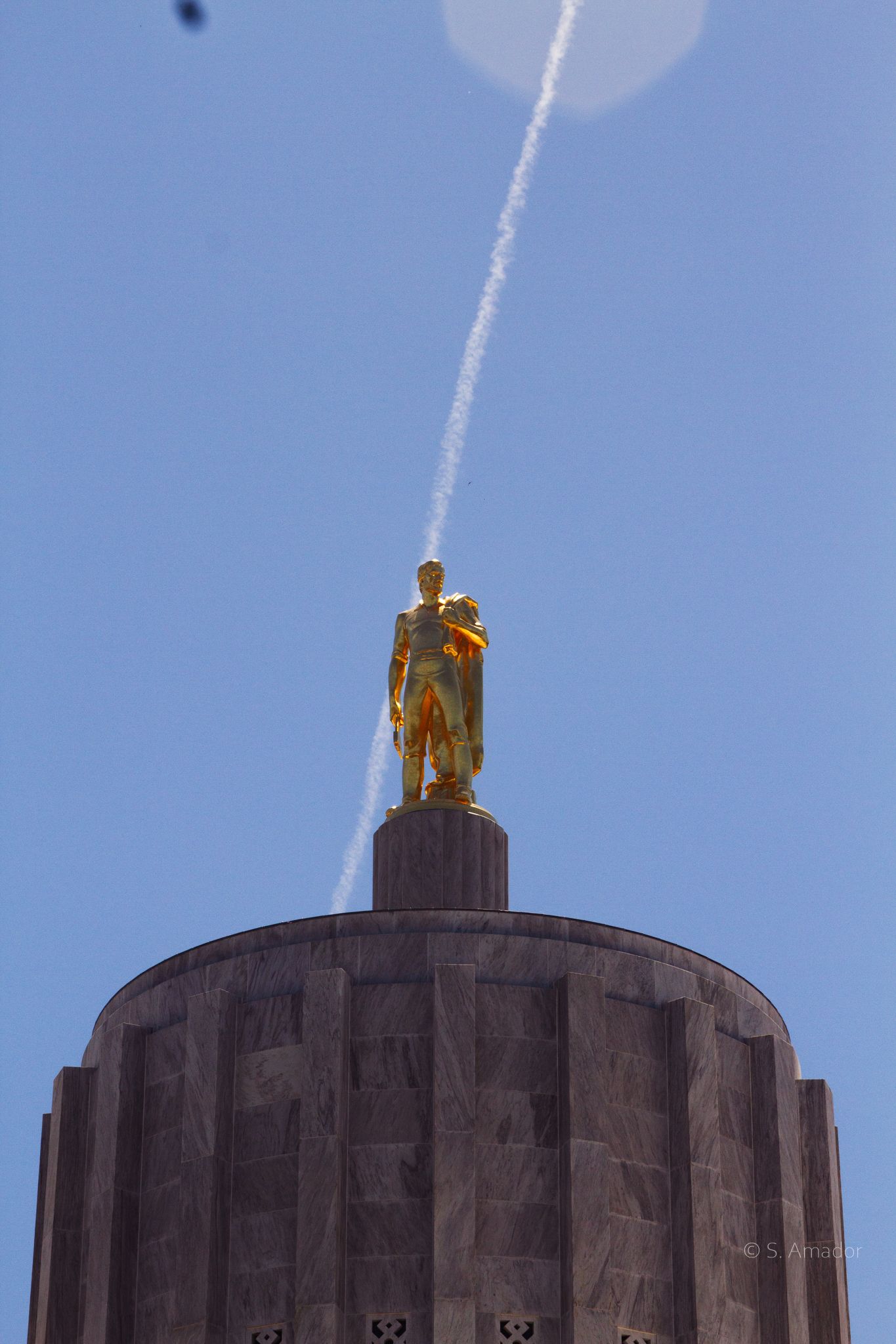 Gold pioneer statue on Oregon Capitol dome with airplane contrail in blue sky.