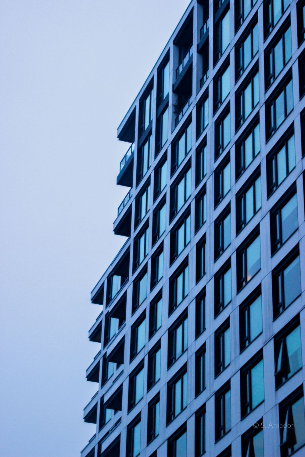 Modern high-rise building facade with blue-tinted glass windows and balconies against overcast sky.