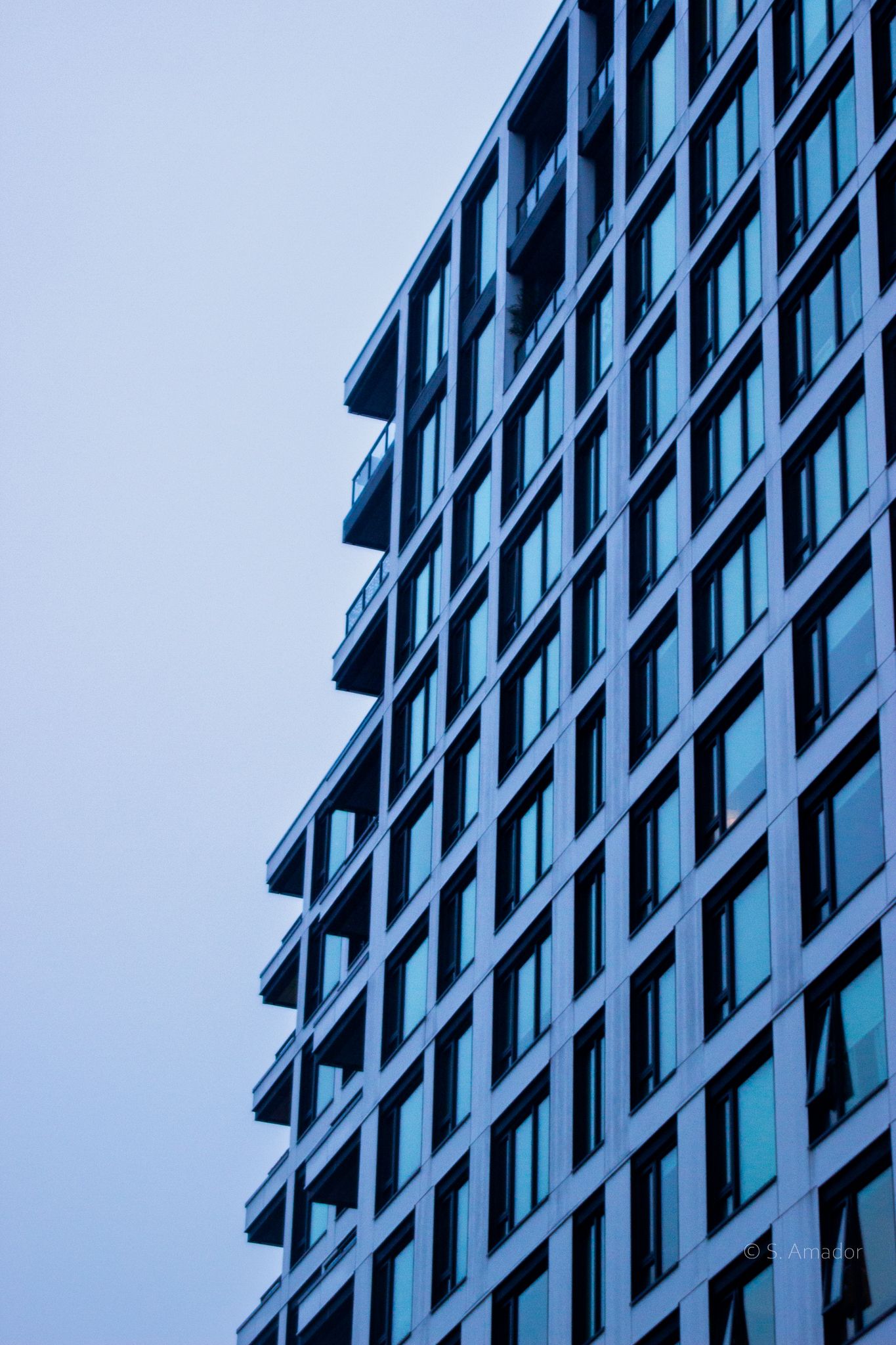 Modern high-rise building facade with blue-tinted glass windows and balconies against overcast sky.