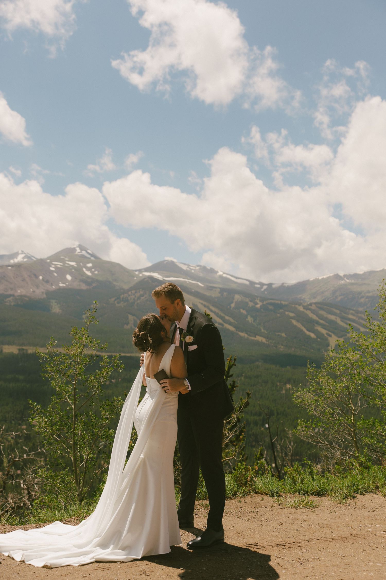 A bride and groom share a kiss on a mountain overlook, with snow-capped peaks and blue skies behind them.