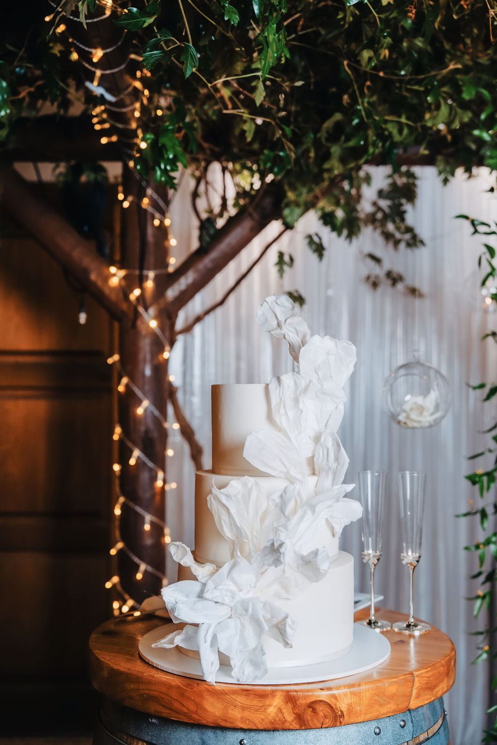 Elegant three-tier white wedding cake with ruffled sugar flowers on a wooden barrel, surrounded by fairy lights and greenery.