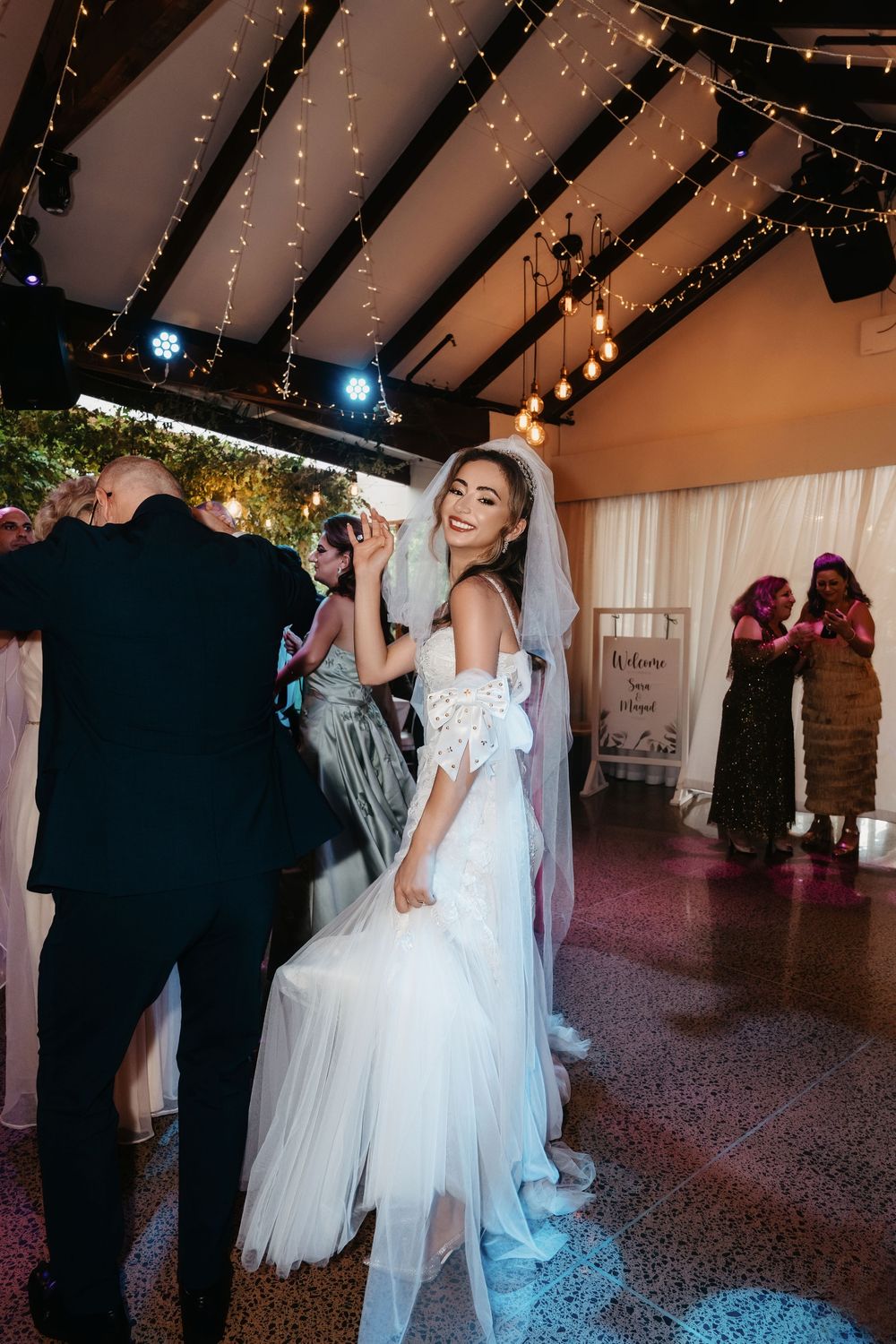 Smiling bride in white lace off-shoulder gown and veil dancing at elegant wedding reception with fairy lights.