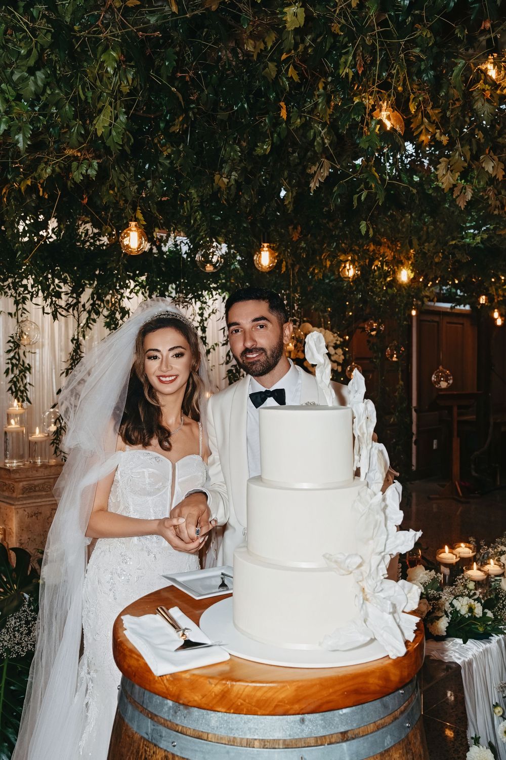 Bride and groom cutting a white four-tier wedding cake on a barrel, surrounded by fairy lights and lush greenery.