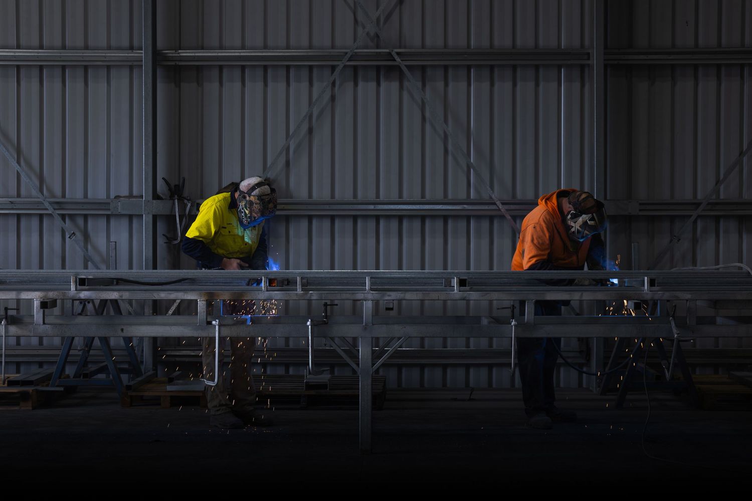 Two welders in safety gear work on metal framework inside an industrial warehouse, with sparks flying.