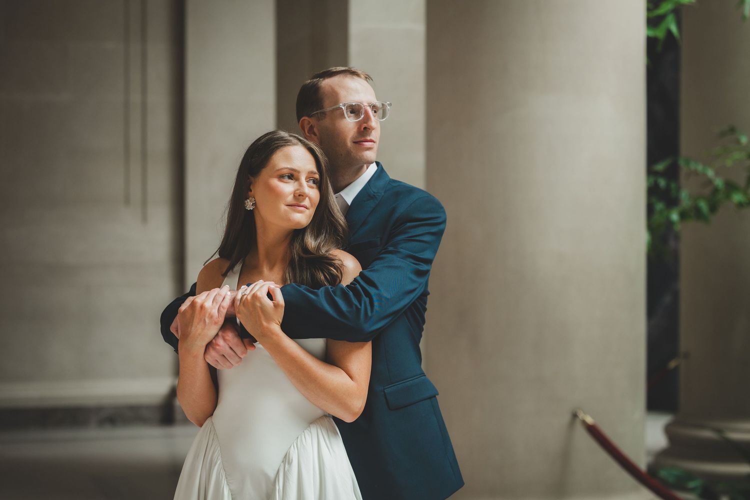 Elegant couple in wedding attire embracing near stone columns, groom in navy suit hugging bride in white dress.