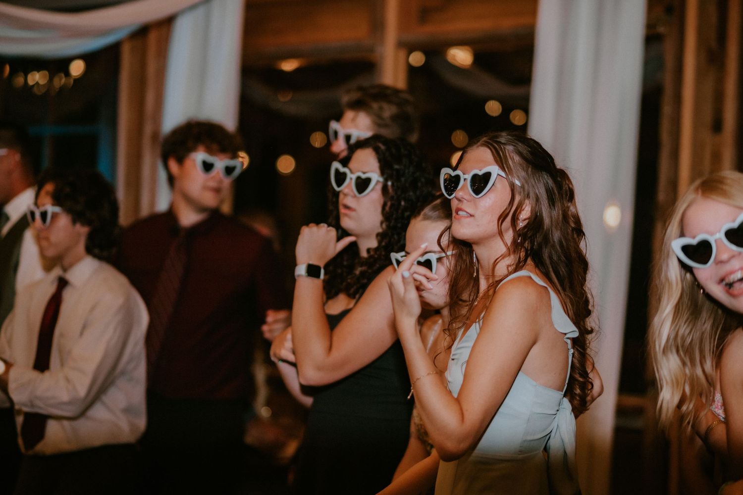 Wedding guests dancing and wearing white heart-shaped sunglasses at a festive indoor reception celebration.