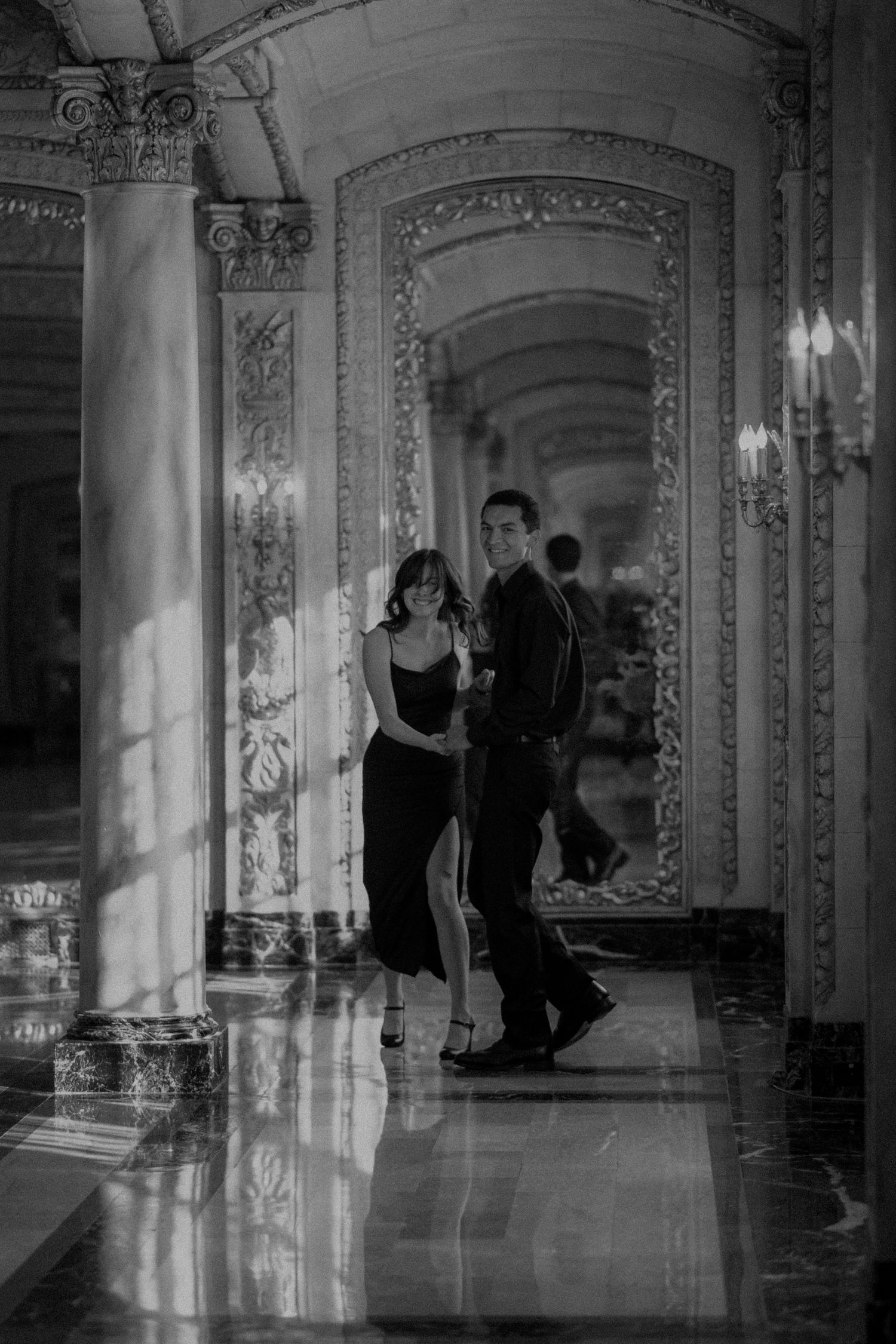 Elegant couple in formal attire posing in a grand ornate hallway with marble columns in black and white.