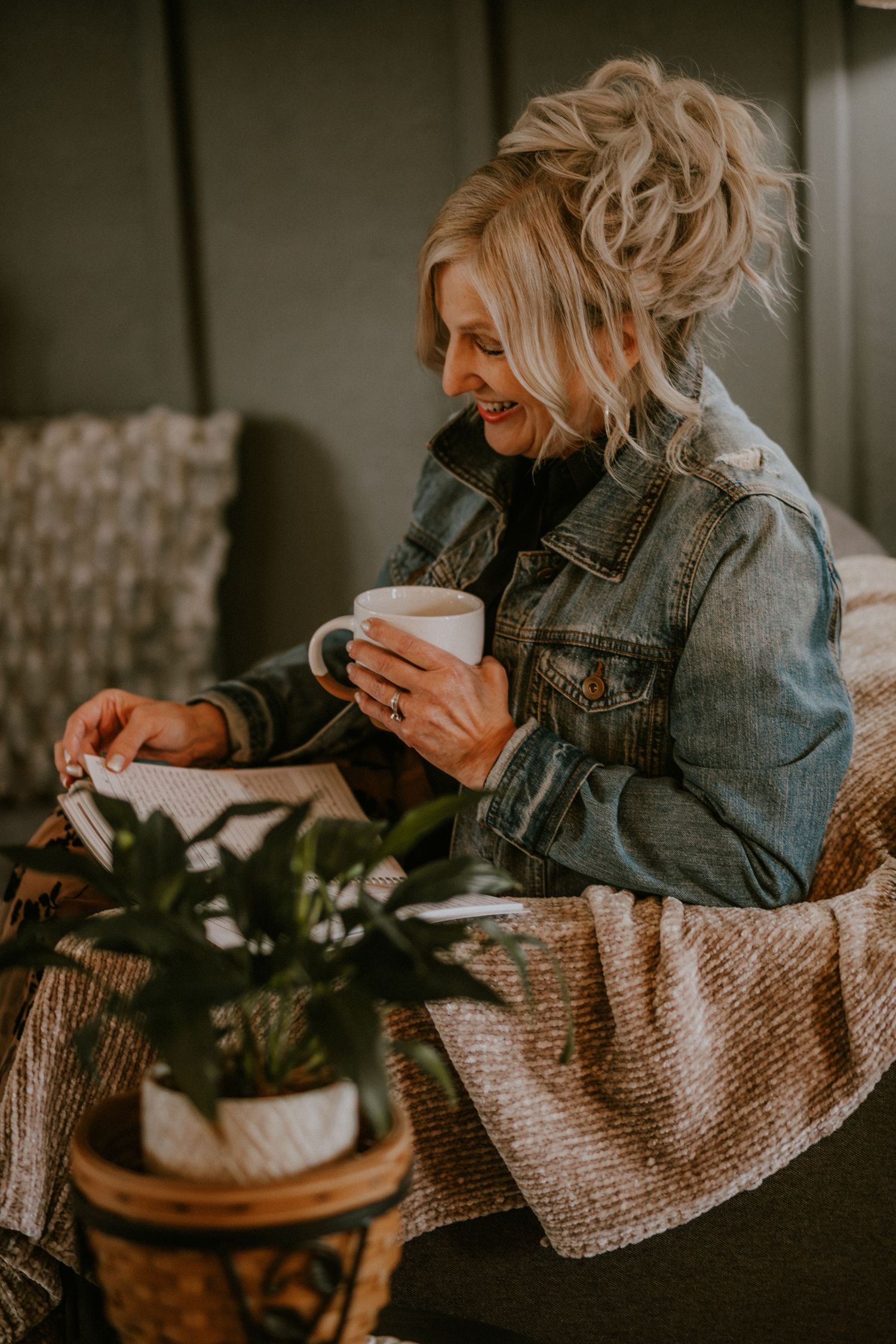 Smiling blonde woman in denim jacket holding white mug while reading a book on a cozy bed with a plant nearby.