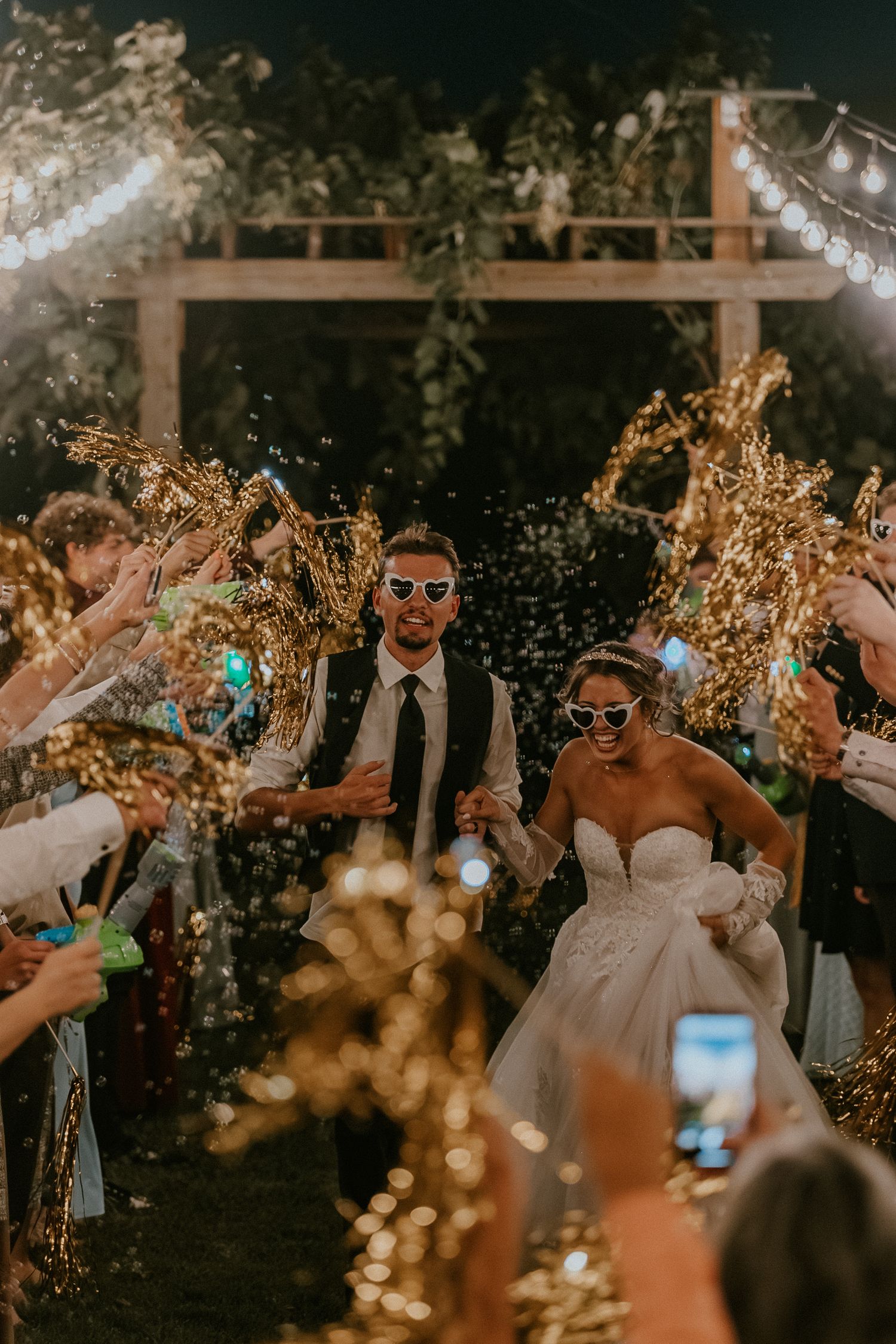 Bride and groom exit wedding reception through golden sparkler tunnel at night, wearing sunglasses, surrounded by cheering guests.