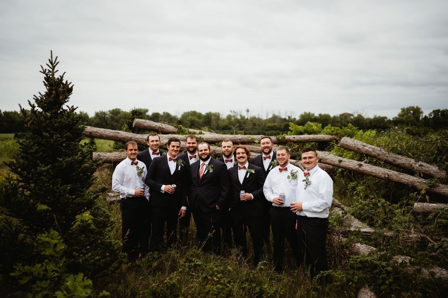 Wedding party celebrating around a rustic tractor outdoors, bridesmaids in coral dresses, surrounded by lush green trees.