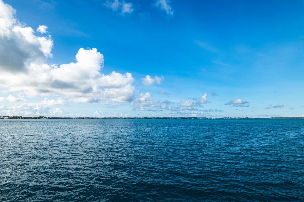 Calm blue ocean under a bright sky with white clouds and distant shoreline on a clear sunny day.